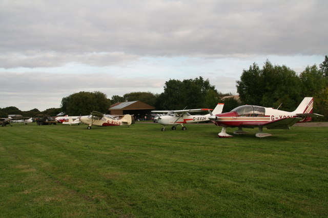 A row of light aircraft on a grass airfield beside a clubhouse